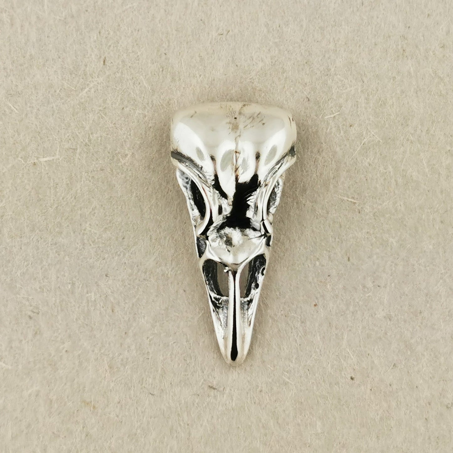 Silver bird skull pendant on a beige background. Sterling silver raven skull charm pendant with gothic and witchy style vibes