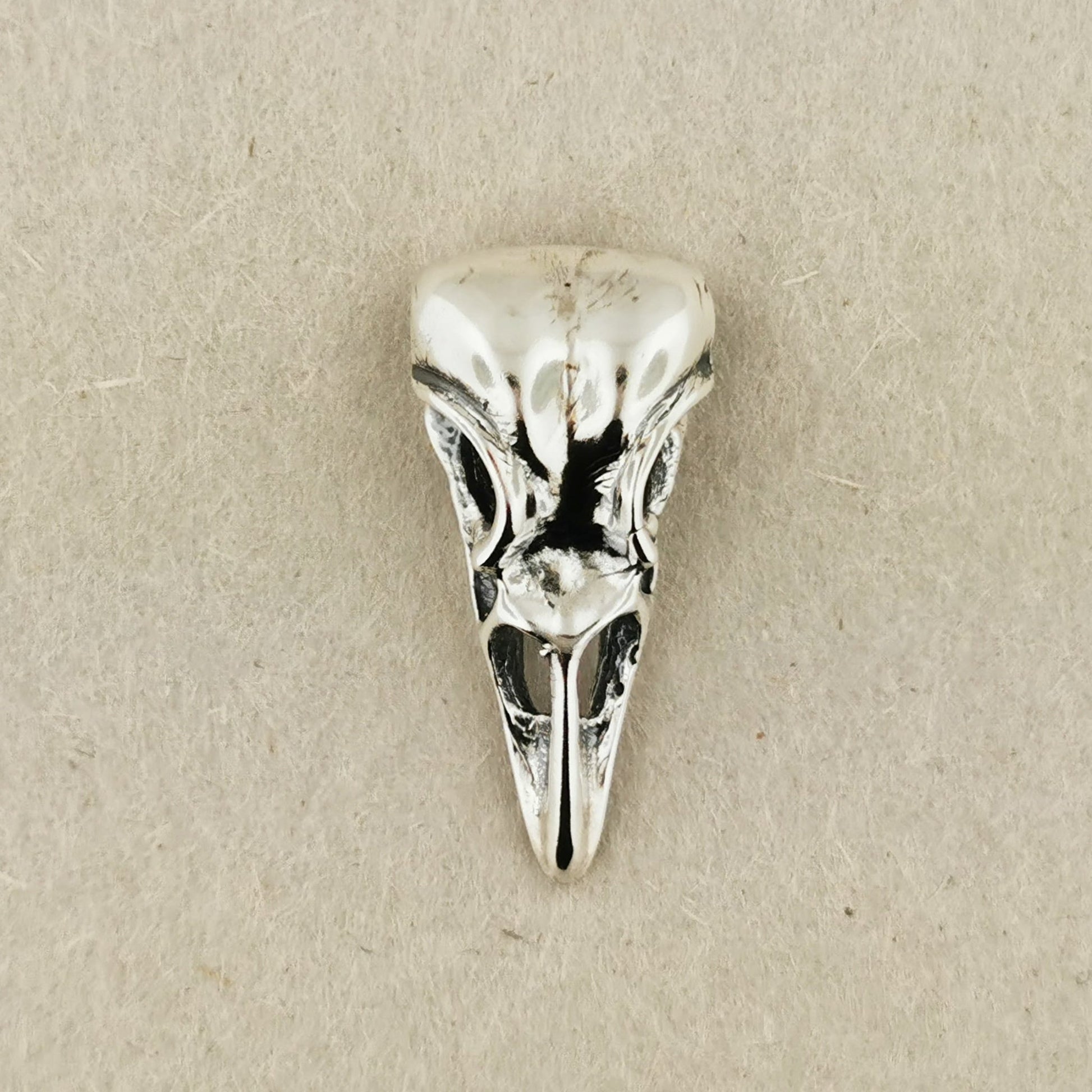 Silver bird skull pendant on a beige background. Sterling silver raven skull charm pendant with gothic and witchy style vibes